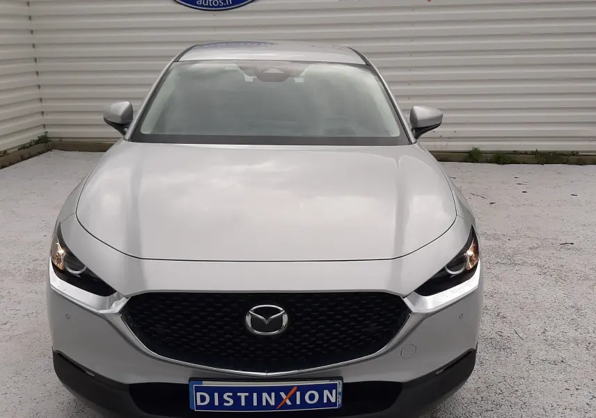 Vue avant d'un Mazda CX-30 blanc céramique 2024 avec calandre noire et phares allumés, stationné devant un mur blanc.