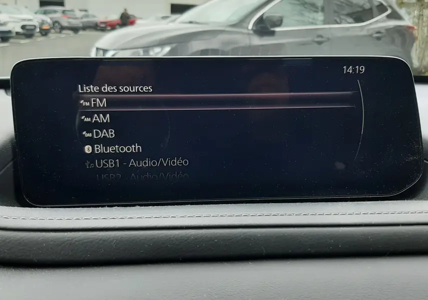 Vue intérieure du tableau de bord du Mazda CX-30 2024 avec écran multimédia affichant les sources audio, arrière gris flou.
