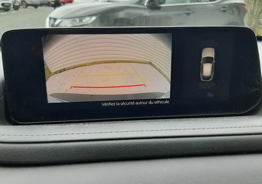 Vue de l'écran intérieur montrant la caméra de recul d'une Mazda CX-30 blanche, avec marquages de stationnement visibles.