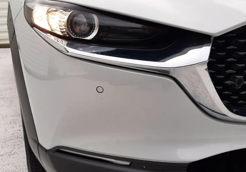 Gros plan sur l'avant gauche du Mazda CX-30 2024 blanc céramique avec phare allumé et calandre chromée.