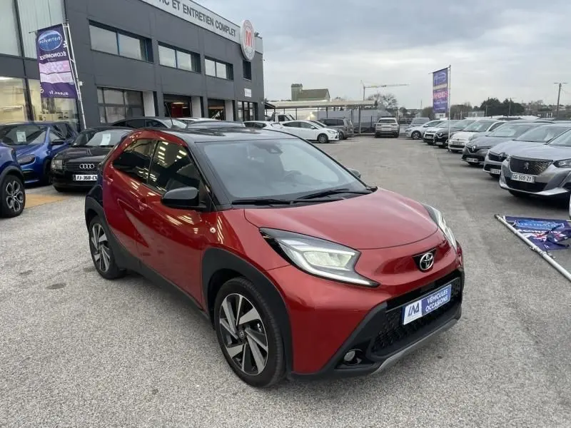 Toyota Aygo X rouge piment avec toit noir, vue 3/4 avant droit, stationnée devant un garage en extérieur.
