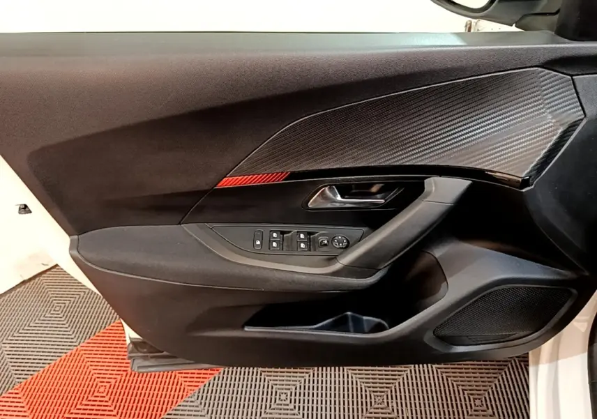 Vue intérieure côté gauche de la porte avant d’une Peugeot 2008 blanche, avec insert imitation carbone et commandes électriques.