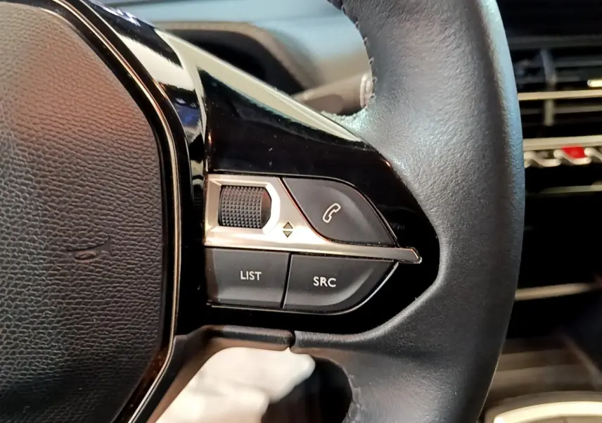 Gros plan sur les commandes gauche du volant cuir noir du Peugeot 2008 blanc, avec boutons multifonctions et molette.