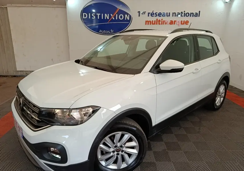 Volkswagen T-Cross blanc vue 3/4 avant droit, avec jantes alliage et barres de toit noires visibles en intérieur showroom.