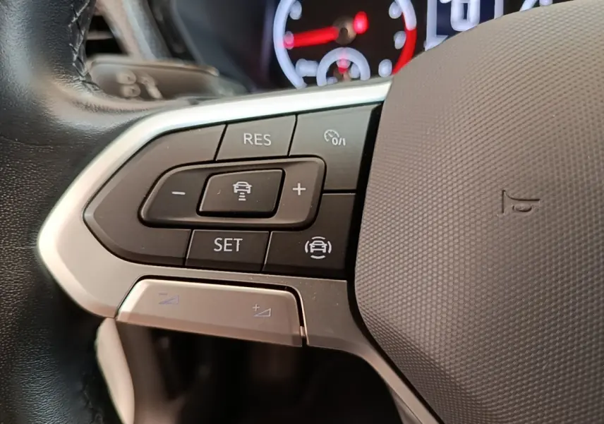 Gros plan sur les commandes de régulateur adaptatif au volant cuir alu du Volkswagen T-Cross blanc, tableau de bord en arrière-plan flou.
