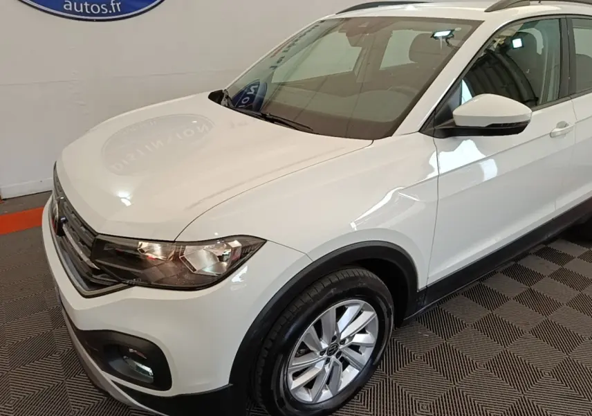 Vue 3/4 avant droit d'un Volkswagen T-Cross blanc avec jantes alliage et barres de toit noires en intérieur showroom.