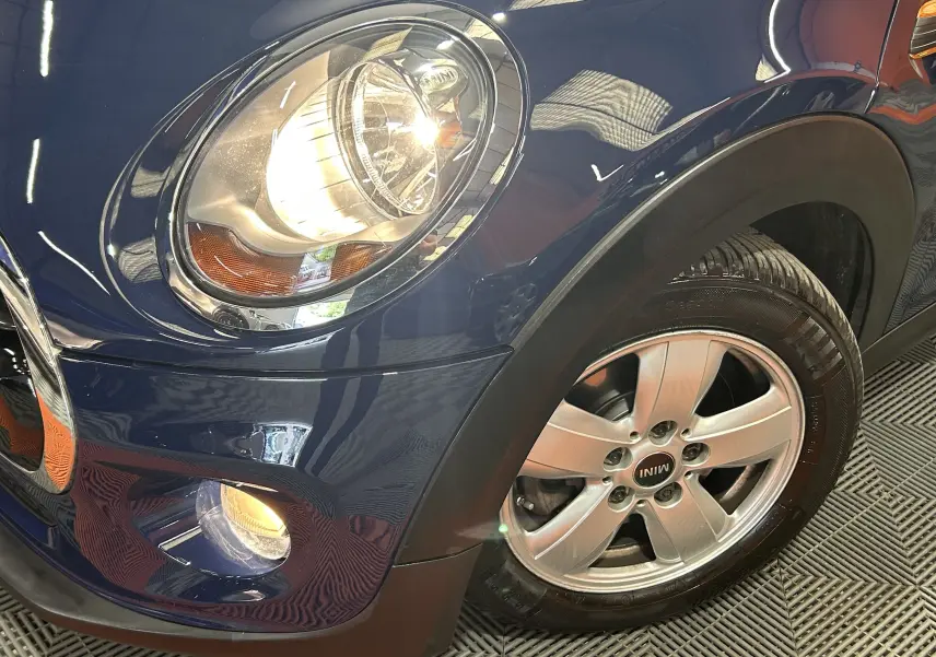 Gros plan sur l'avant droit bleu d'une MINI 5 portes avec phare allumé et jante alu siglée MINI.