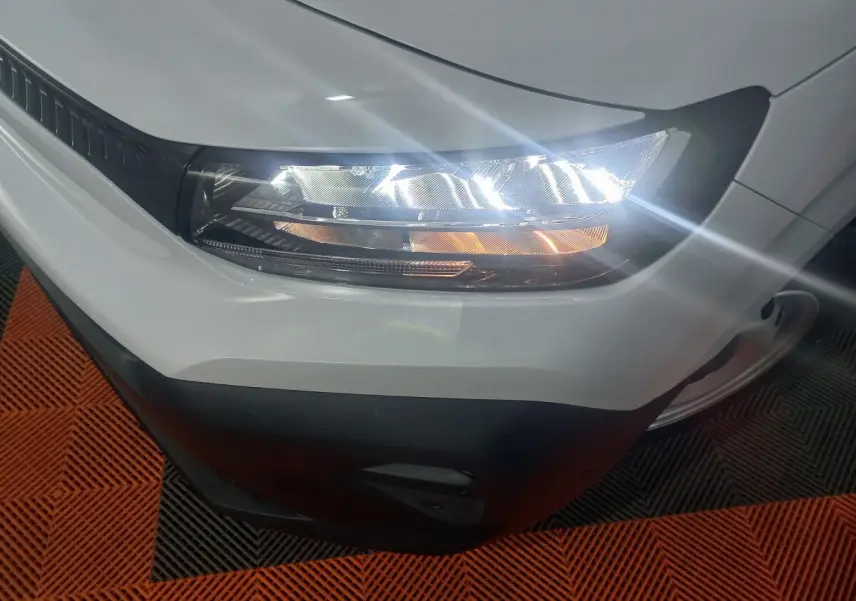 Gros plan sur le phare avant droit allumé d’un Citroën Berlingo blanc, avec pare-chocs noir visible.