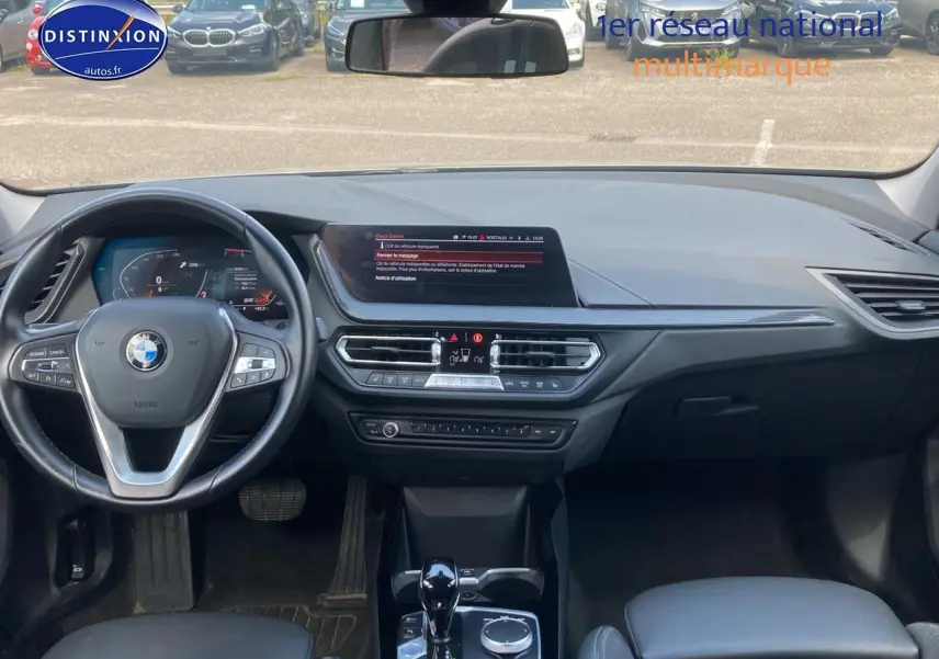 Vue intérieure avant de la BMW Série 1 118i 2023, tableau de bord noir avec volant multifonction et écran central.