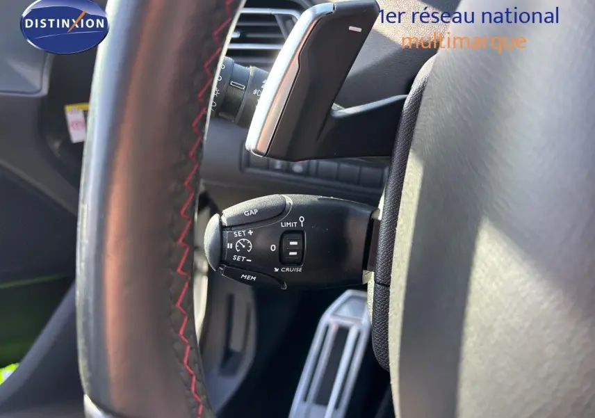 Gros plan sur la commande de régulateur de vitesse derrière le volant cuir noir avec surpiqûres rouges d'une Peugeot 308 blanche nacrée.