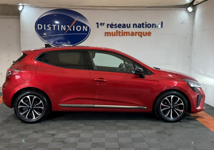 Profil droit d'une Renault Clio Techno TCe 90 rouge 2024, avec jantes alliage et feux LED visibles en intérieur showroom.