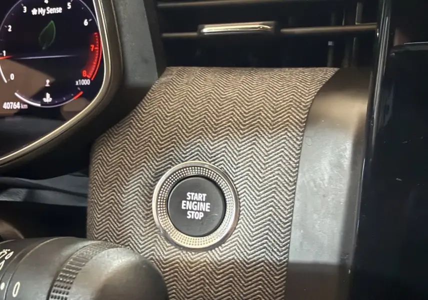 Gros plan sur le bouton Start Engine Stop entouré d’un habillage gris à motif chevrons, avec le combiné d’instruments partiellement visible.