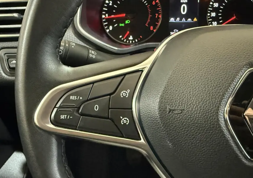 Gros plan sur le volant cuir synthétique noir de la Renault Clio avec commandes de régulateur de vitesse et tableau de bord éclairé.