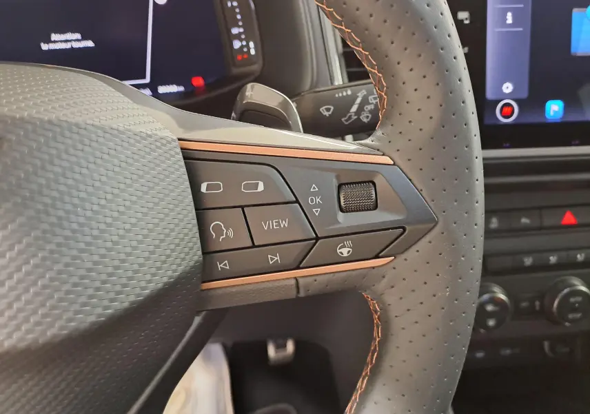 Gros plan sur les commandes au volant côté gauche du CUPRA Ateca 2024 noir magique avec surpiqûres orange et écran digital en arrière-plan.