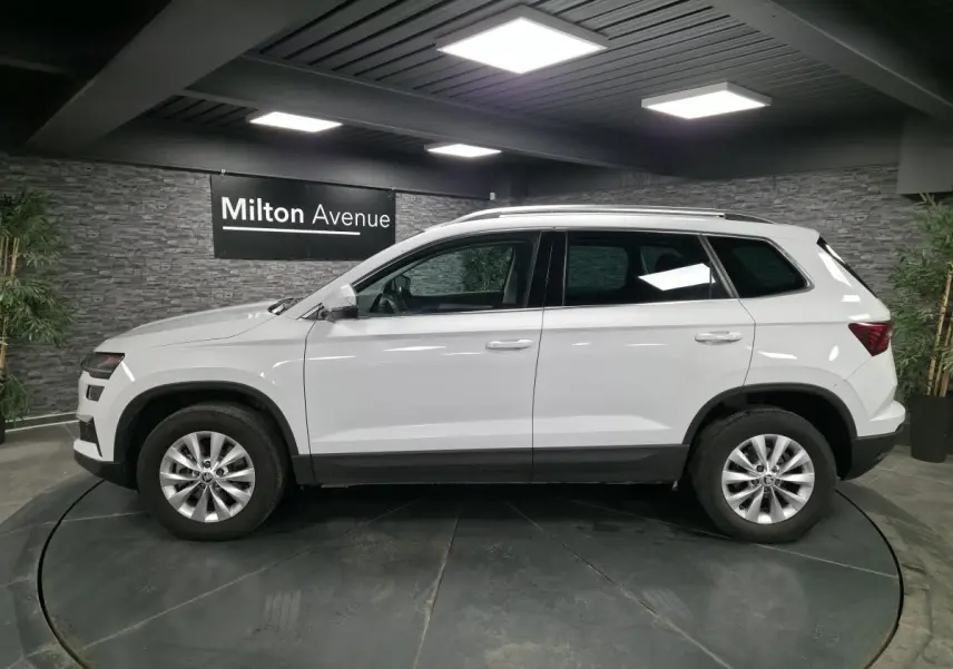 Vue de profil côté gauche d'un SUV Skoda Karoq blanc 2024 avec jantes alliage et barres de toit dans un showroom.
