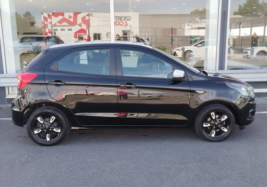 Vue de profil côté gauche d'une Ford KA+ noire avec toit blanc et jantes bi-ton, garée devant une vitrine.