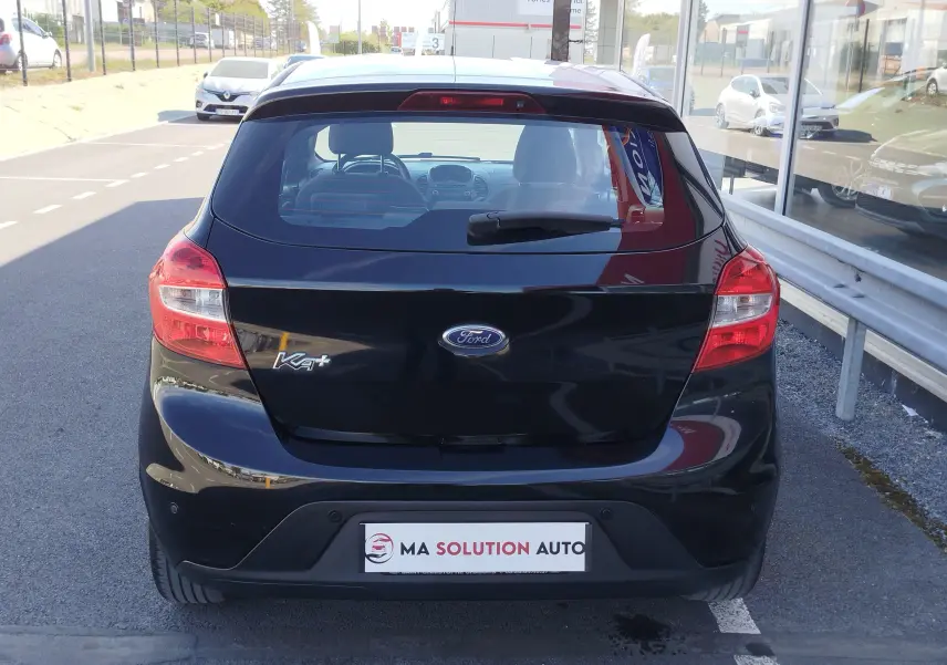 Vue arrière d'une Ford KA+ noire Shadow avec toit blanc, stationnée devant un bâtiment vitré.