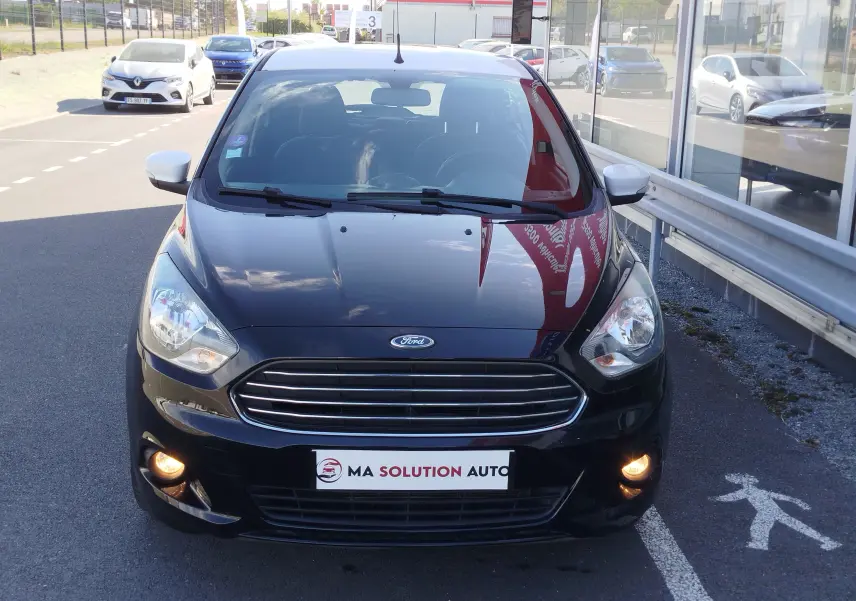 Vue avant d'une Ford KA+ noire avec toit blanc, phares allumés, stationnée devant un bâtiment vitré.