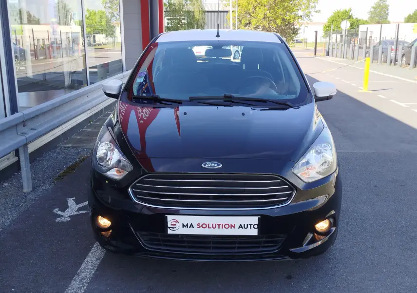 Vue avant d'une Ford KA+ noire avec toit blanc, phares et feux antibrouillard allumés, stationnée devant un bâtiment.