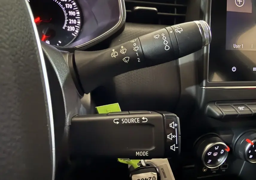 Gros plan sur la commande multifonction noire à gauche du volant d’une Renault Clio Business blanche, tableau de bord partiel visible.