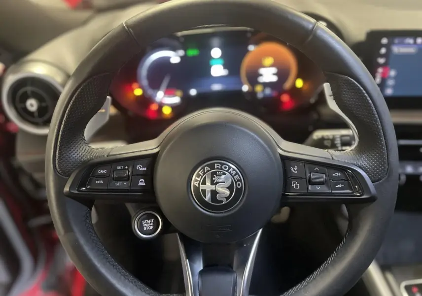 Volant sport cuir noir Alfa Romeo avec commandes intégrées et bouton start/stop, tableau de bord digital flou en arrière-plan.