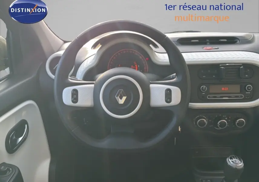 Vue intérieure centrée sur le volant noir et blanc de la Renault Twingo gris acier, avec tableau de bord et levier de vitesses manuelle visibles.
