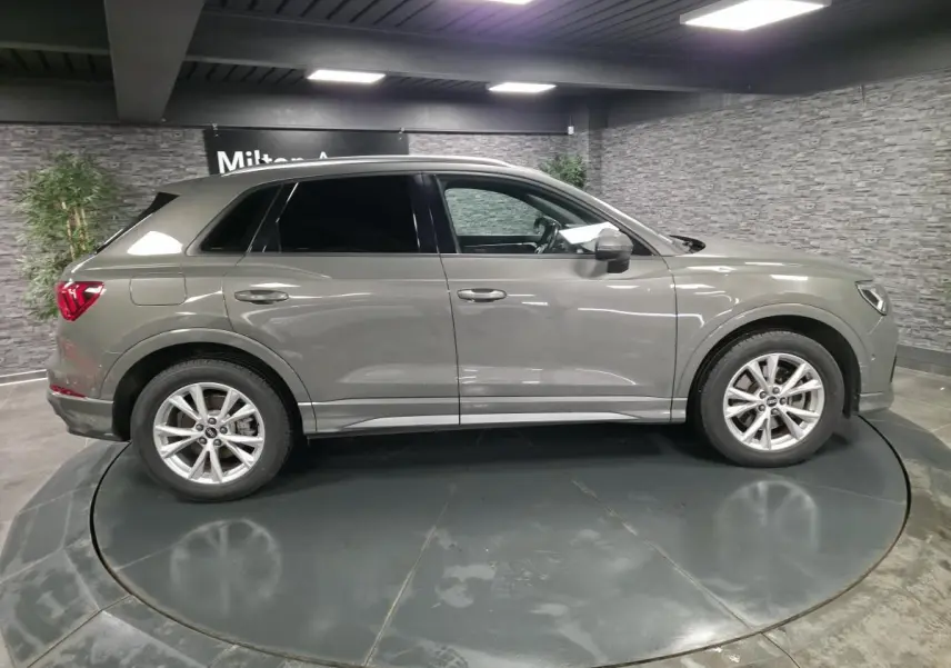 Audi Q3 gris en vue profil côté gauche, vitres surteintées et jantes alliage 5 branches en V visibles en intérieur showroom.
