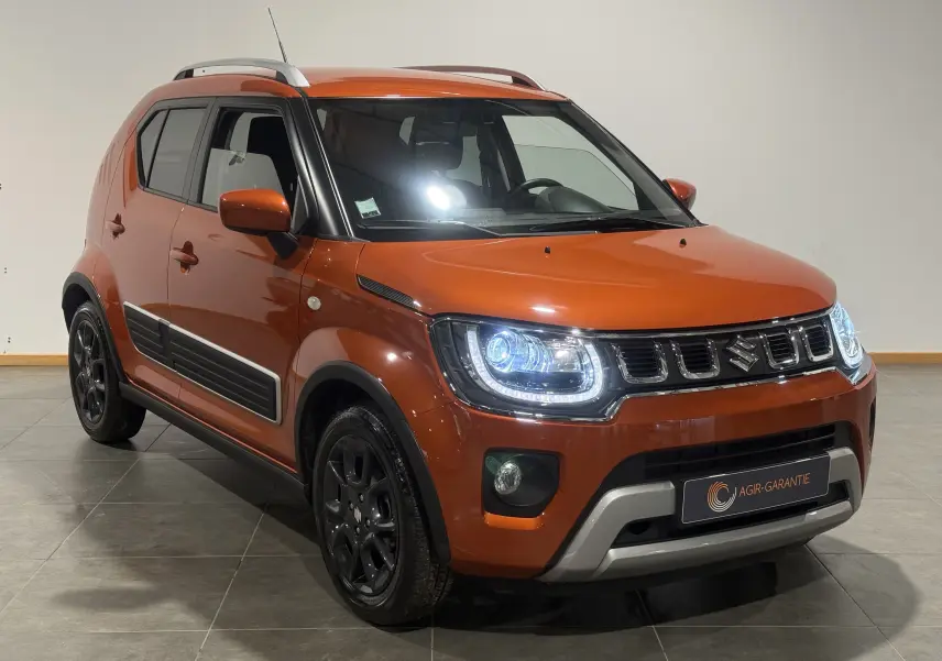 Suzuki Ignis orange vue 3/4 avant droit avec phares allumés et protections latérales noires visibles.
