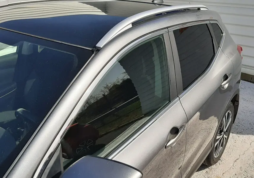 Vue 3/4 avant droit du Nissan Qashqai marron métal 2018, avec barres de toit argentées et vitres teintées.