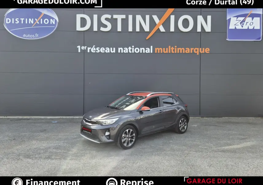 Vue 3/4 avant droite d'un Kia Stonic gris avec toit et rétroviseurs orange devant un mur gris.