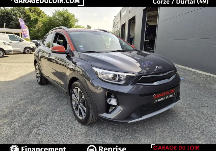 Vue 3/4 avant d’un KIA STONIC gris avec rétroviseurs et montants orange, stationné devant un garage.