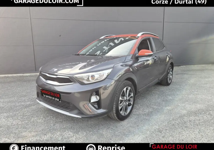 Vue 3/4 avant d'un KIA Stonic gris avec toit et rétroviseurs orange, sur fond urbain neutre.