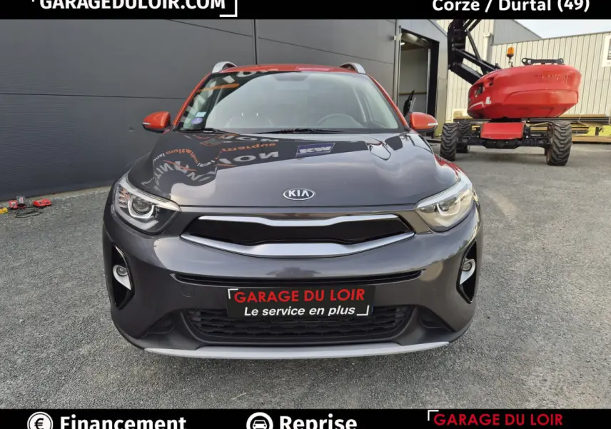Vue avant d'un KIA Stonic 1.0 T-GDi 100 ACTIVE gris avec rétroviseurs orange, stationné devant un bâtiment industriel.