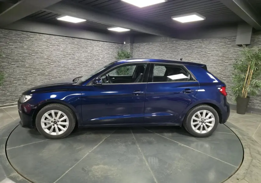 Audi A1 Sportback bleu métallisé vue de profil côté gauche dans un showroom avec sol gris et murs en pierre.