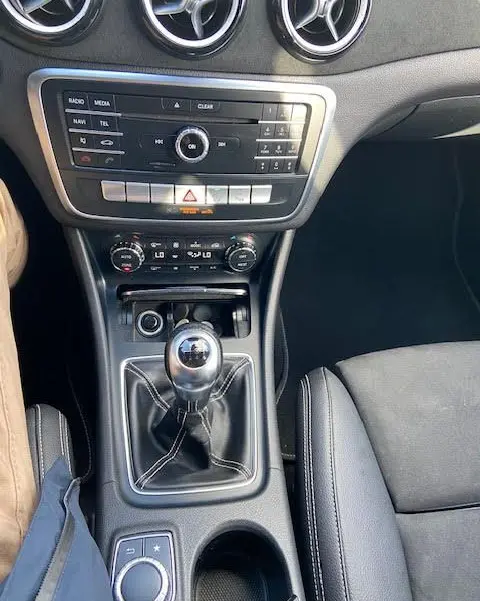 Vue intérieure centrée sur la console centrale d'une Mercedes CLA Shooting Brake 200D blanche, avec levier de vitesses manuel et aérateurs ronds chromés.