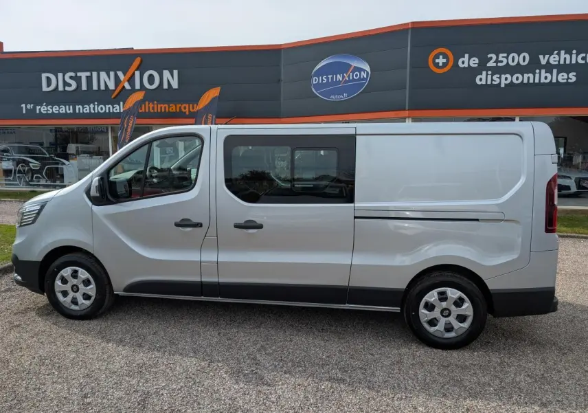 Profil côté gauche d’un Renault Trafic Fourgon gris avec cabine approfondie et enjoliveurs 16 pouces, stationné devant un concessionnaire.