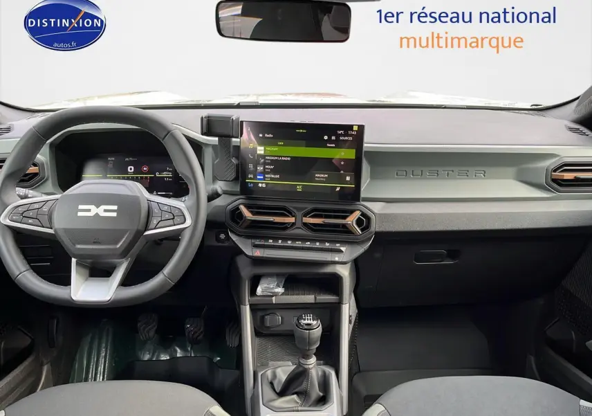 Vue intérieure avant du Dacia Duster 2025 avec tableau de bord gris et écran tactile central, volant multifonction et boîte manuelle.