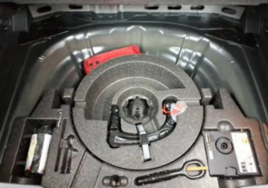 Vue plongeante sur le coffre ouvert d’un Volkswagen T-Roc noir, montrant le rangement pour roue de secours et outils.