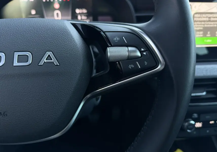 Gros plan sur le volant cuir noir multifonctions de la Skoda Scala TSI 116, avec commandes intégrées et tableau de bord numérique flou.