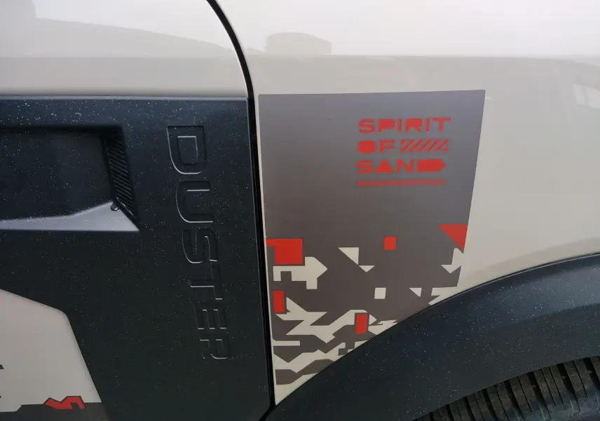 Gros plan sur l'aile avant droite beige sandstone du Dacia Duster Spirit of Sand avec logo et marquage décoratif rouge et gris.