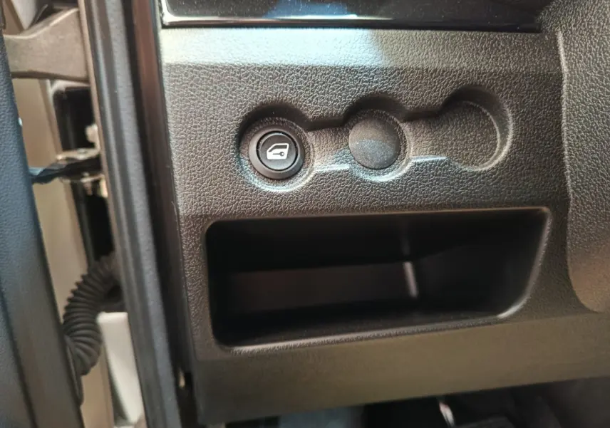 Détail intérieur montrant le bouton d'ouverture du coffre sur la partie inférieure gauche du tableau de bord d'une Aixam Ambition blanche.