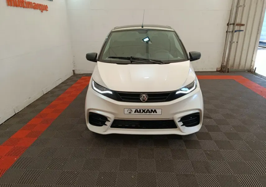 Vue avant d'une Aixam Ambition 0.5 City Coupe Sport blanche avec phares allumés et rétroviseurs noirs en intérieur showroom.