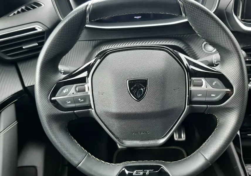 Vue rapprochée du volant cuir noir perforé avec logo Peugeot et sigle GT, tableau de bord et commandes visibles.