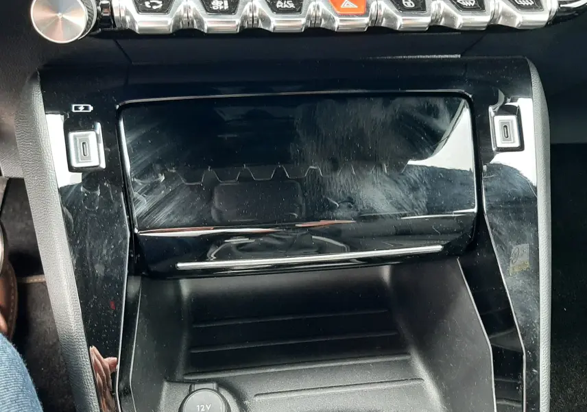 Vue rapprochée de la console centrale noire brillante de la Peugeot 208 1.2 100CH GT, avec boutons chromés et prise 12V visible.
