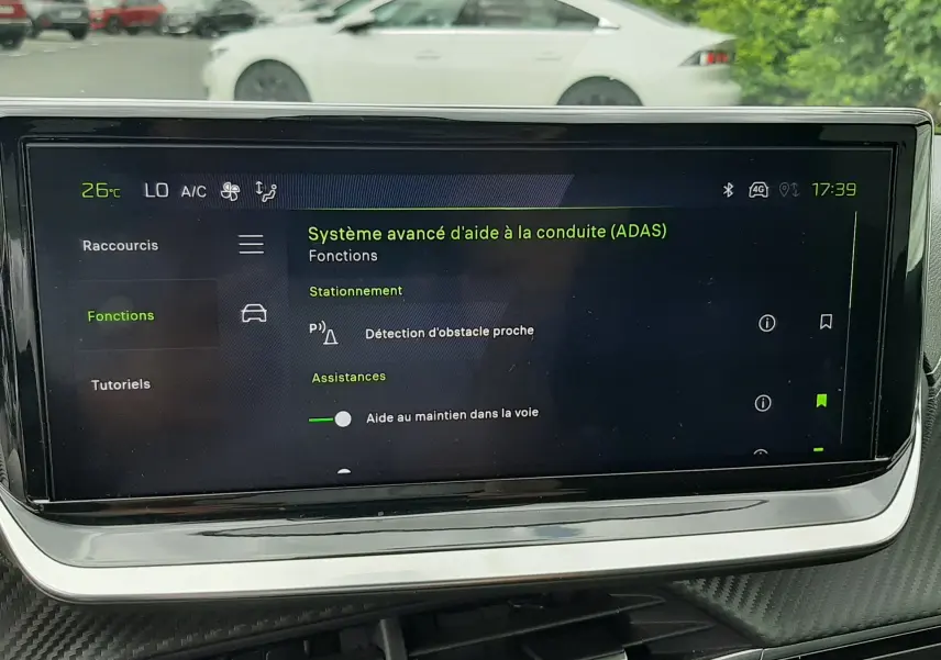 Vue intérieure centrée sur l'écran tactile affichant l'aide à la conduite, avec une Peugeot 208 gris Artense visible en arrière-plan côté gauche.