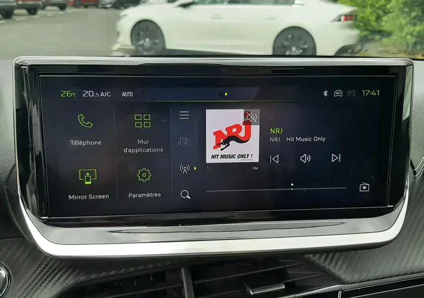 Vue intérieure centrée sur l'écran tactile multimédia de la Peugeot 208 GT, avec une Peugeot 208 gris Artense visible en arrière-plan.