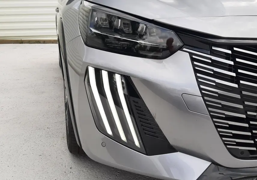 Gros plan sur l’avant droit d’une Peugeot 208 gris Artense, mettant en valeur les phares LED et la calandre distinctive.