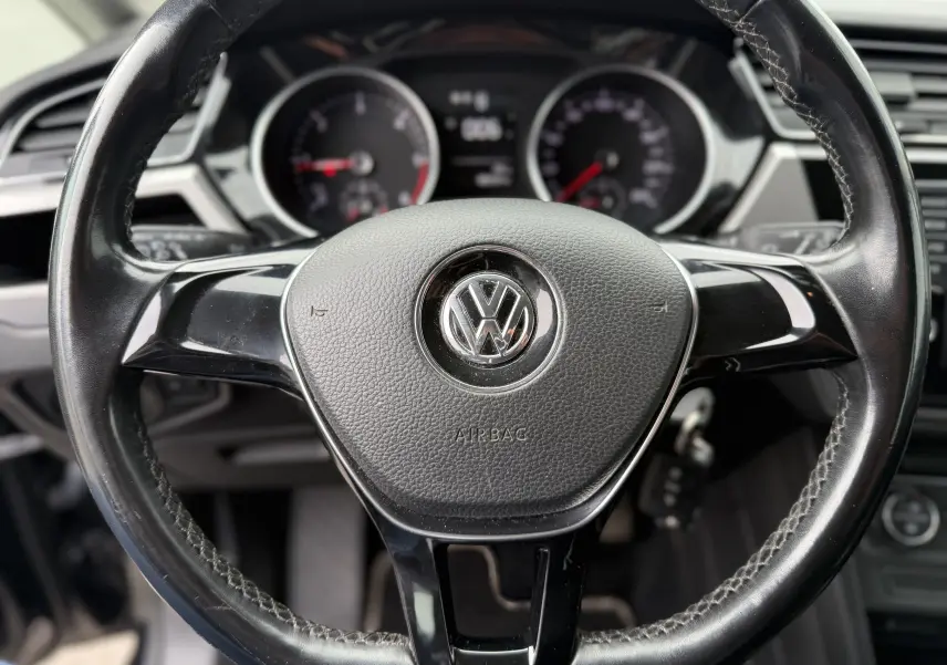Gros plan sur le volant noir du Volkswagen Touran 2017, avec logo VW centré et tableau de bord flou en arrière-plan.