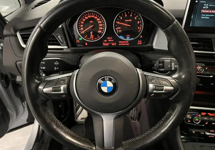 Vue intérieure centrée sur le volant M cuir noir de la BMW Série 2 225xe Active Tourer avec tableau de bord et écran multimédia allumés.