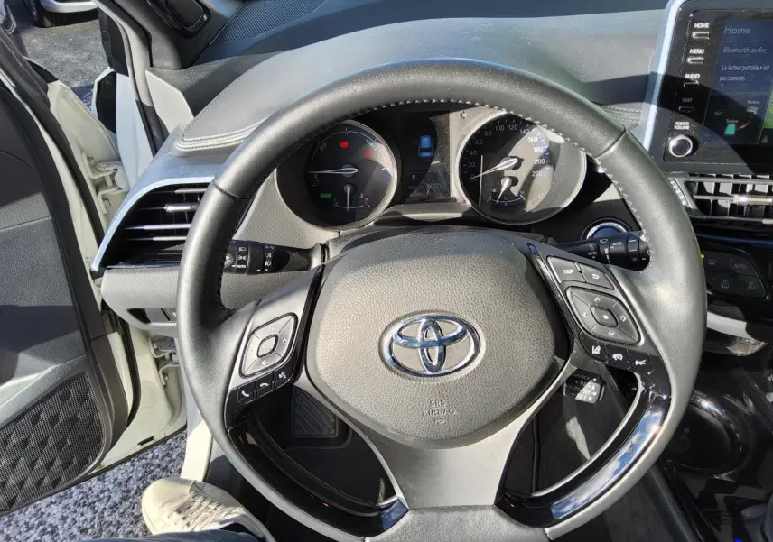 Vue intérieure centrée sur le volant noir du Toyota C-HR blanc nacré, avec tableau de bord et écran tactile visibles.